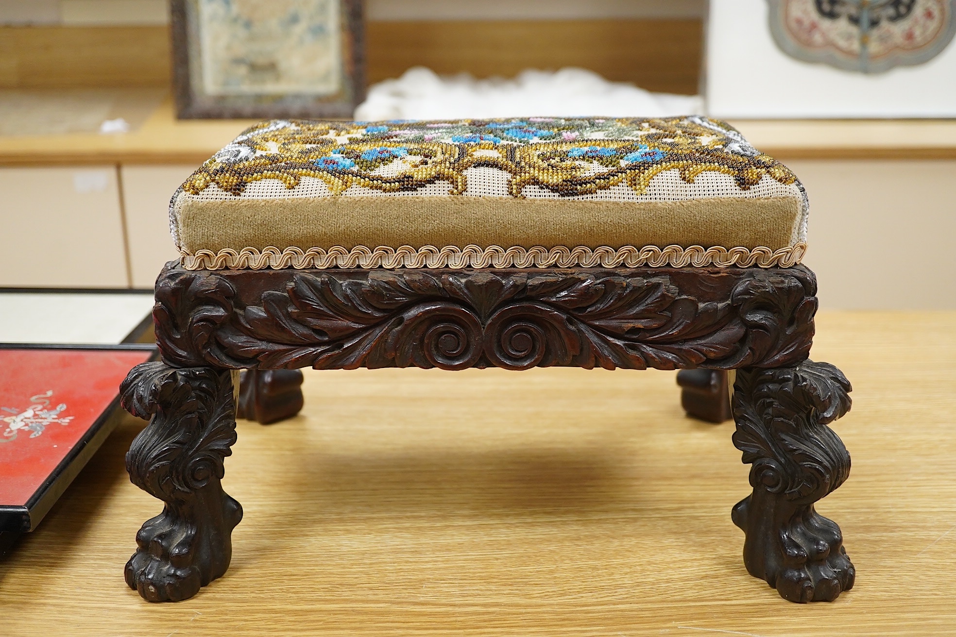 A 19th century ornately carved paw foot, Berlin beadwork upholstered footstool, the bead work a scrolling multi-coloured floral pattern, upholstered into a heavily carved frame and legs on carved paw feet, beadwork 21cm